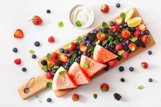A wooden serving board with sliced watermelon, assorted berries, mint leaves, lime wedges, and a small bowl of white yogurt on a white surface.