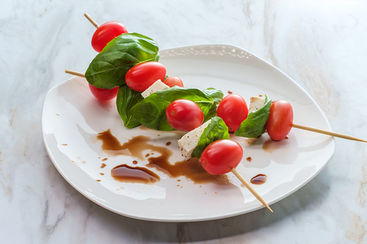 Caprese salad (tomatoes, mozzarella and basil) threaded on wooden skewers and drizzled with balsamic dressing resting on a white plate