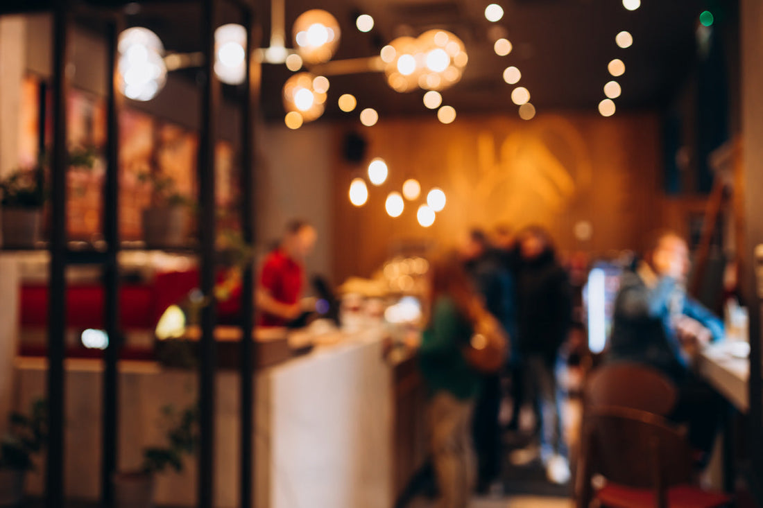 A blurry image of a bustling cafe captures people at the counter and seated, enjoying a moment away. Warm lighting and modern decor provide a cozy atmosphere, perfect for those mindful of nutrition while dining out.