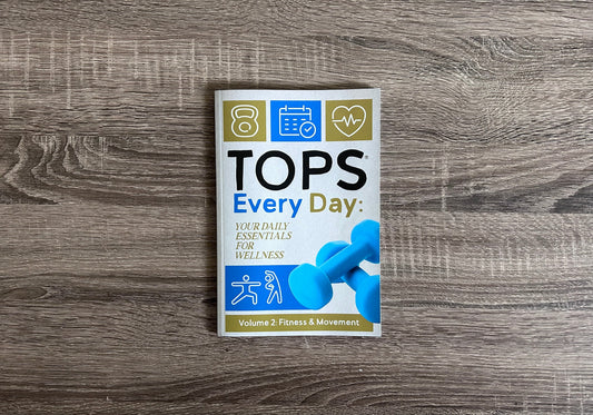 A cover of a booklet resting on a wooden background with the title "TOPS Every Day, Volume 2:Fitness & Movement"