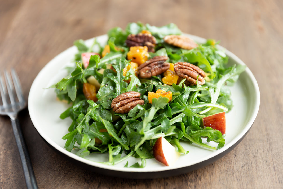 A fresh salad on a white plate featuring arugula, sliced apples, butternut squash cubes and toasted pecans