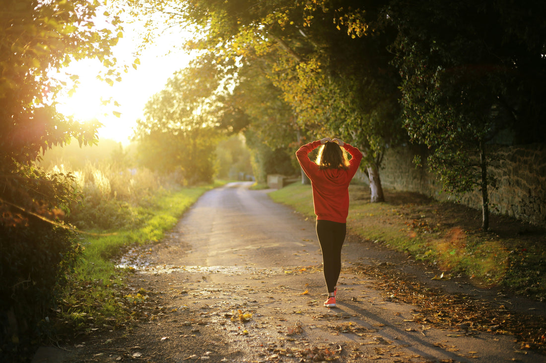 A person in a red hoodie and black leggings walks away down a leaf-strewn country lane, hands resting on their head, as golden sunlight streams through the autumn trees.