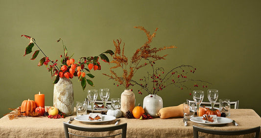 Elegant Thanksgiving table setting with autumn centerpiece featuring red berries in ceramic vases, mini pumpkins, candles, butternut squash, and neutral dinnerware against sage green wall