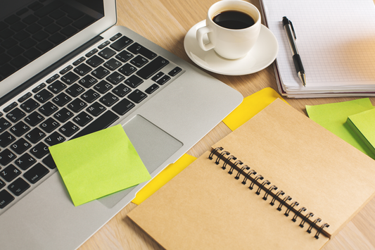 A cup of espresso next to a laptop with a sticky note and open notebook